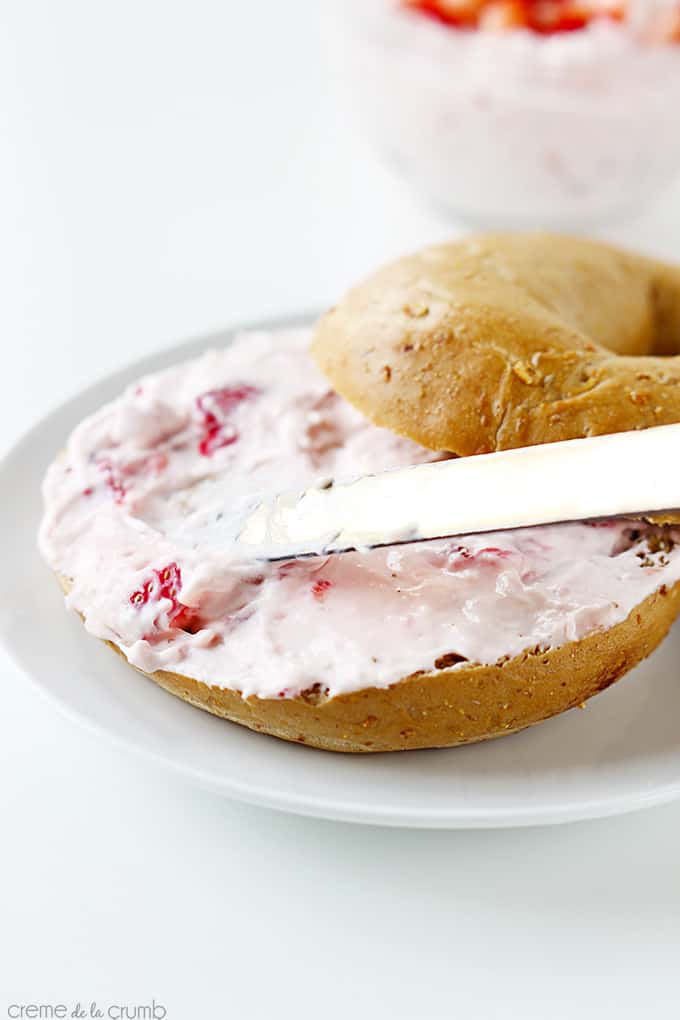 a knife spreading strawberry cream cheese shmear on a half of a bagel with the other half on the side.