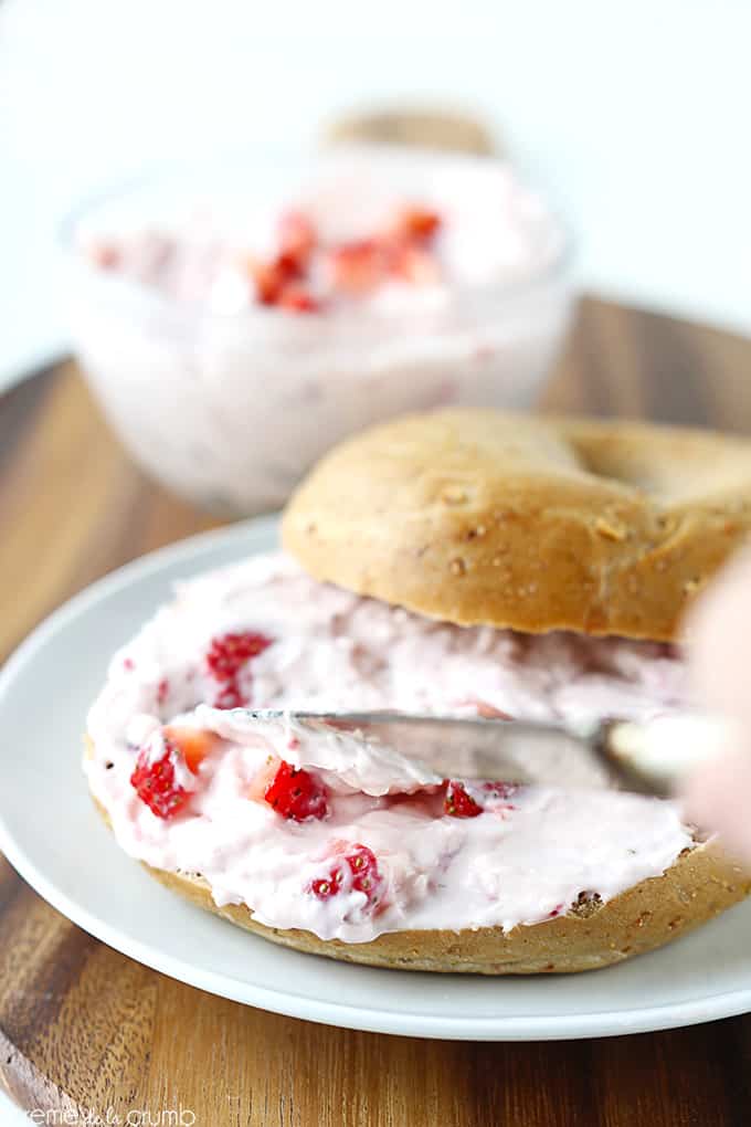 a knife spreading strawberry cream cheese shmear on a bagel on a plate with more shmear in a bowl faded in the background all on a wooden cutting board.