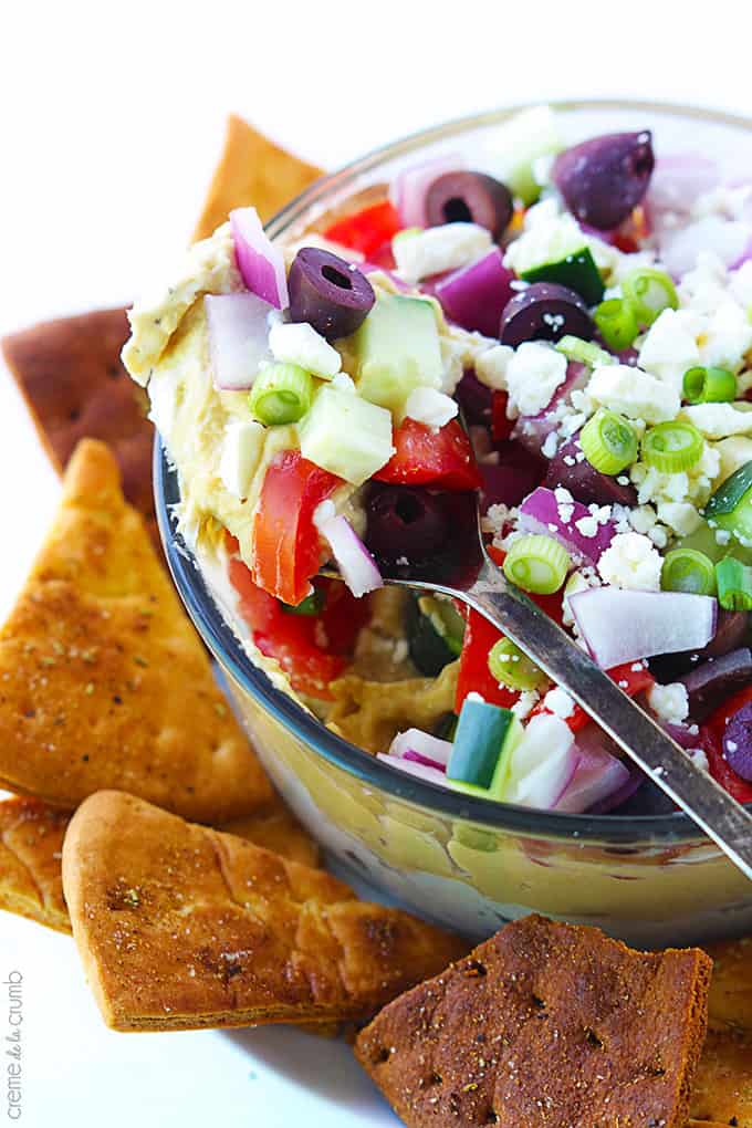 top view of 7 layer Greek hummus in a bowl with pita chips on the side with a spoon with a scoop of hummus being lifted from the bowl.