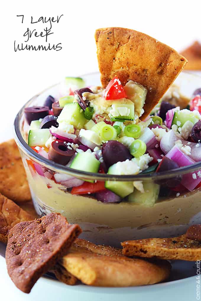 7 layer Greek hummus in a bowl with a pita chip in the bowl and more chips on the side with the title of the recipe written on the top left corner of the image.