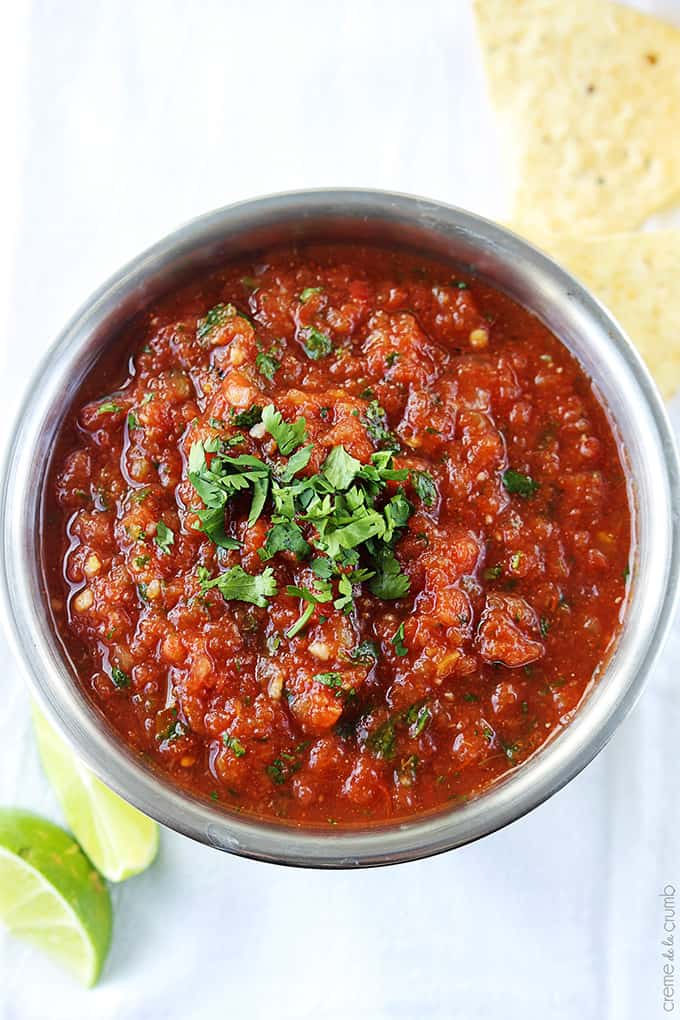 close up of blender salsa in a metal bowl with slices of limes and chips on the side.