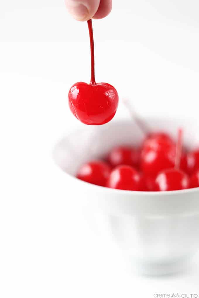 fingers holding a cherry with more cherries in a bowl faded in the background.
