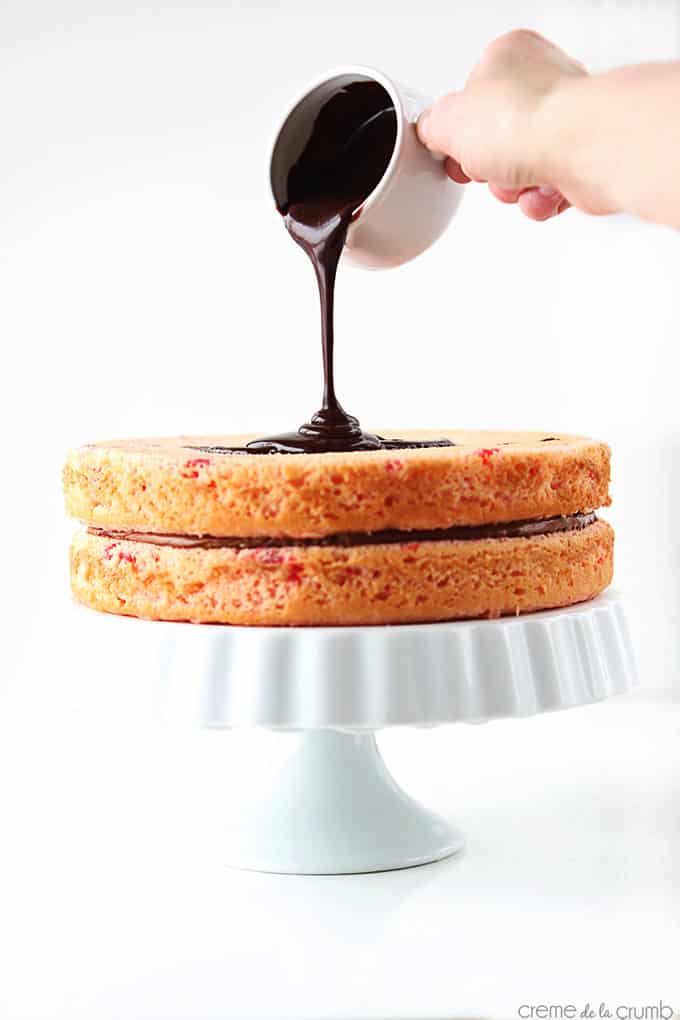 two layers of cherry chip cake on a cake stand with chocolate ganache frosting being poured on the second layer.