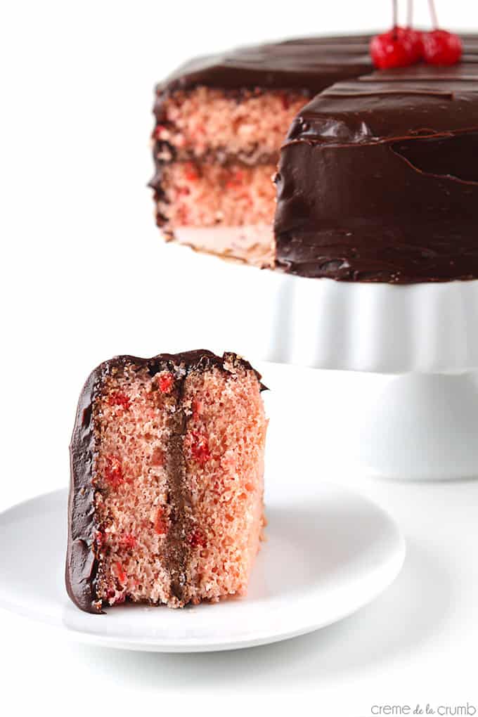 a piece of cherry chip cake with chocolate ganache frosting on a plate with the rest of the cake in the background on a cake stand.