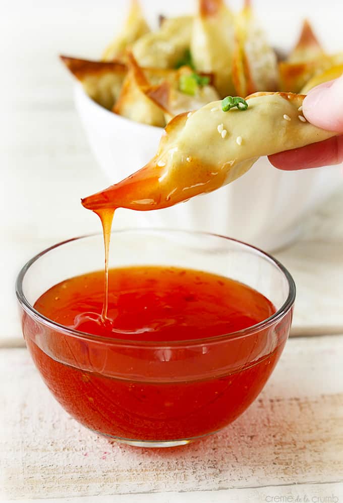 a hand holding a oven baked crab avocado wonton just dipped in sweet and sour sauce with more wontons in a bowl faded in the background.