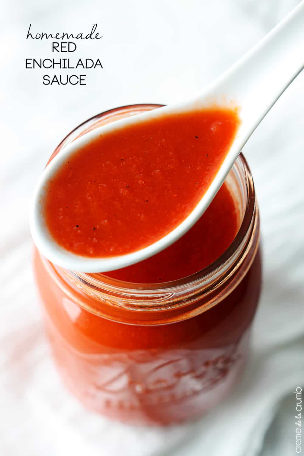 top view of a spoon full of homemade red enchilada sauce on top of a mason jar with sauce in it with the title of the recipe written on the top left corner of the image.