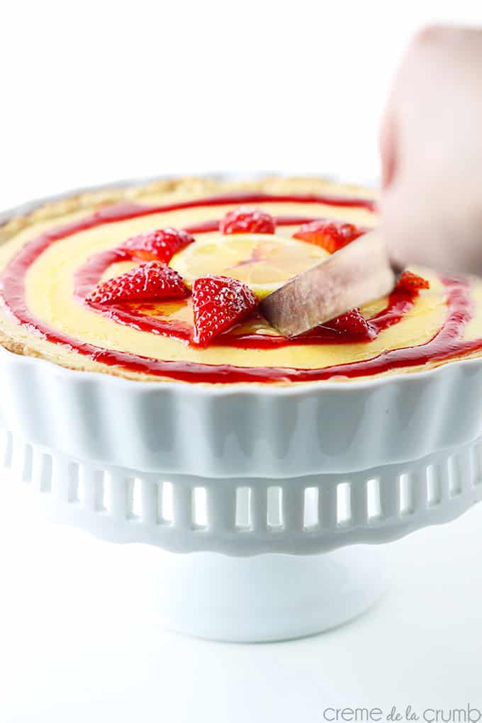 a hand holding a knife cutting into honey lemon & strawberry tart in a pedestal dish.