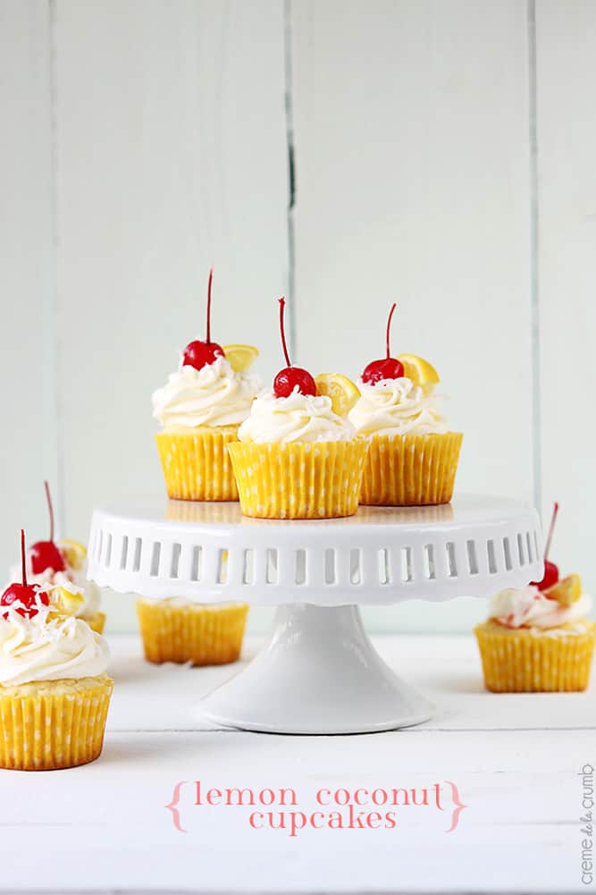 lemon coconut cupcakes topped with cherries and slices of lemon on a cake pedestal tray and on the table with the title of the recipe written on the bottom of the image.