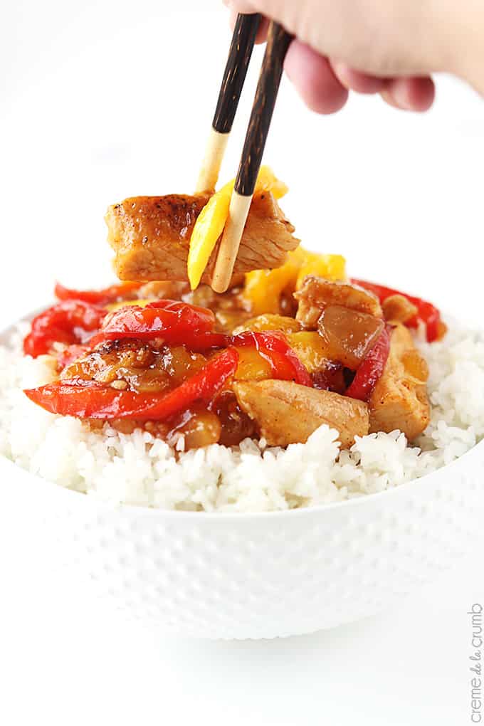 mango chicken stir fry on rice in a bowl with a hand holding chopsticks grabbing a piece of chicken.