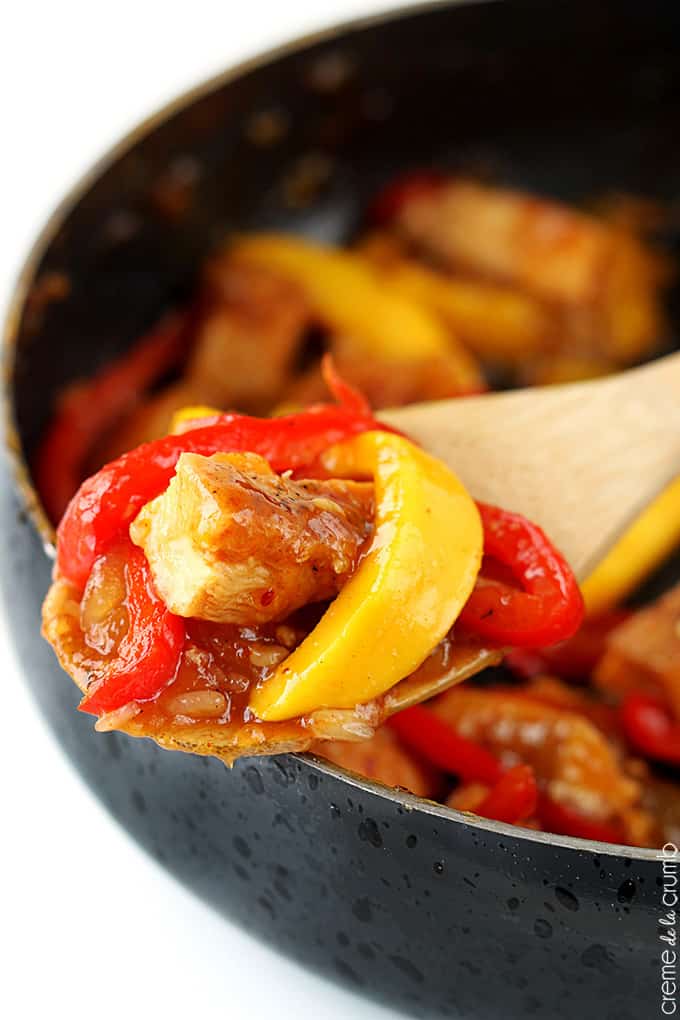a wooden serving spoon with a scoop of mango chicken stir fry on it with more chicken in a skillet faded in the background.