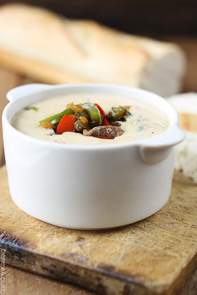 Philly cheesesteak dip in a bowl on a wooden cutting board with a loaf of French bread faded in the background.