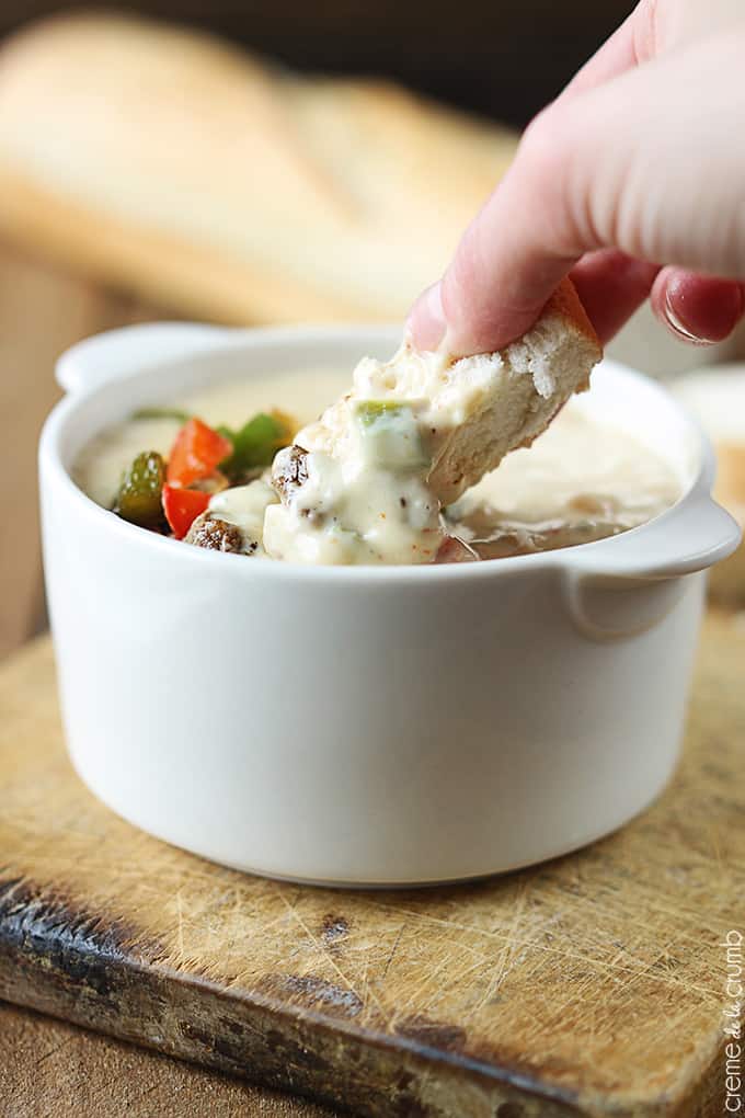 a hand dipping a piece of bread in Philly cheesesteak dip in a bowl on a wooden cutting board with a loaf of French bread faded in the background.