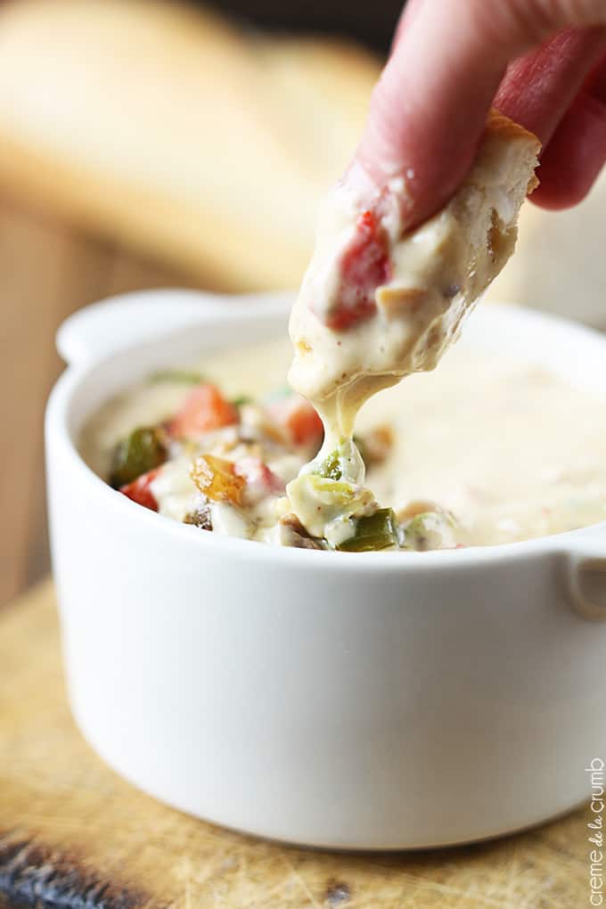 a hand holding a piece of bread just dipped in Philly cheesesteak dip in a bowl.