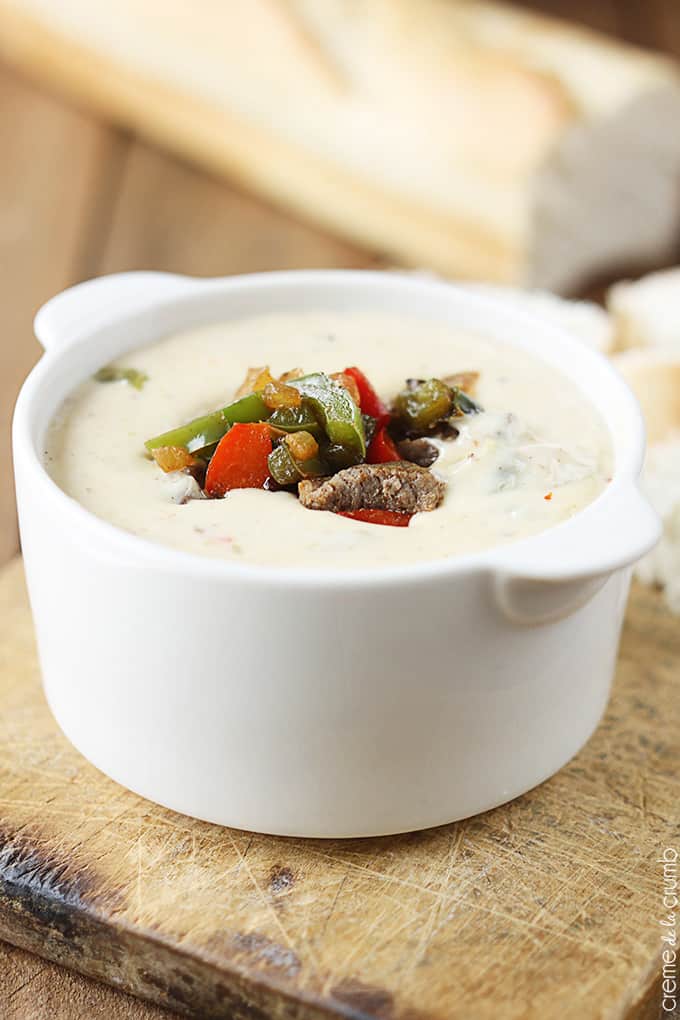 Philly cheesesteak dip in a bowl on a cutting board with a loaf of French bread faded in the background.