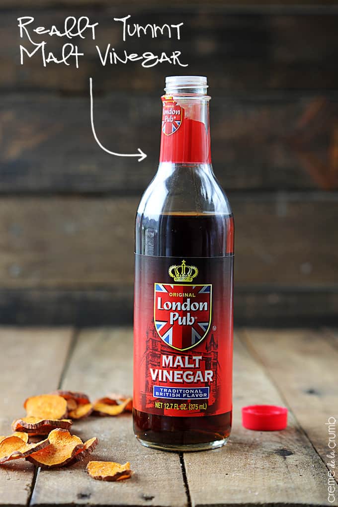 a bottle of malt vinegar with chips on the side on a wooden table with the name of the vinegar written on the top left corner of the image with an arrow pointing to the bottle.