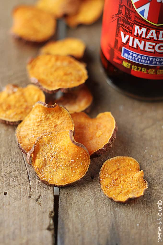 close up of salt & vinegar sweet potato chips with a bottle of malt vinegar on a wooden table.