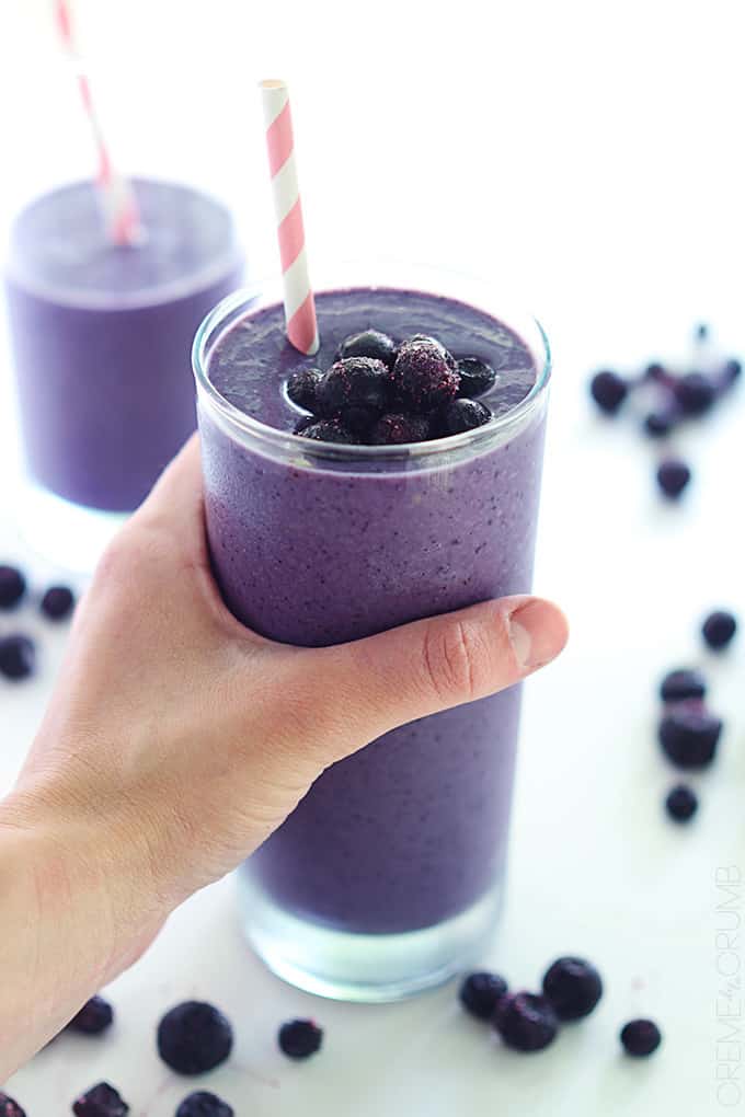 a hand holding a blueberry smoothie in a glass topped with berries and a straw with another smoothie and more berries faded in the background.