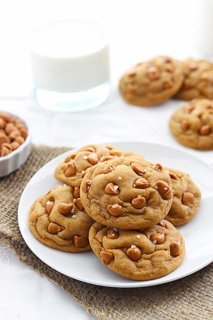 butterscotch chip pudding cookies on a plate with more cookies, butterscotch chips and a glass of milk on the side.