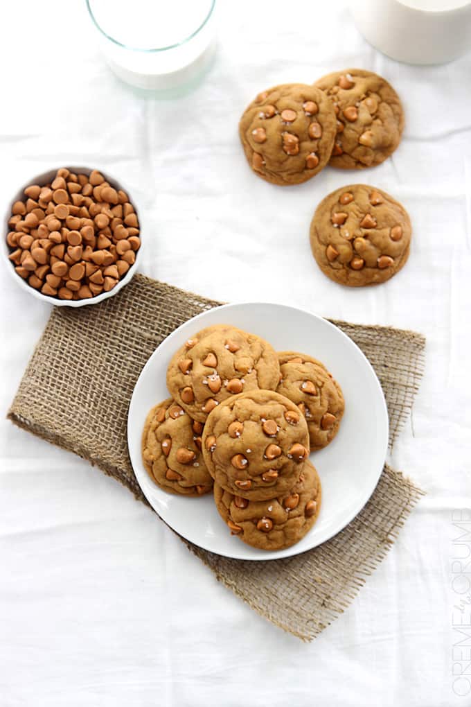 top view of butterscotch chip pudding cookies on a plate with more cookies, butterscotch chips, and milk on the side.