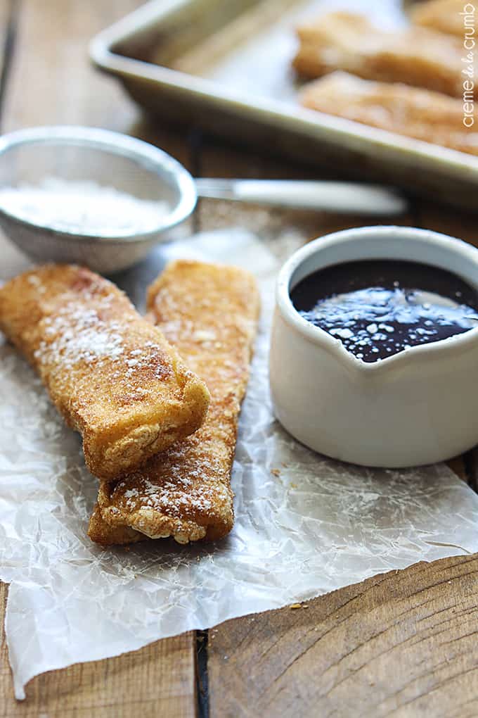 two stacked cream cheese stuffed churros next to chocolate sauce with powdered sugar in a sifter faded in the background.