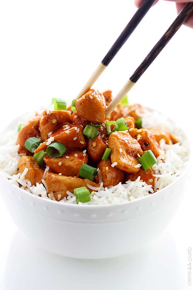 a hand holding chopsticks taking a piece of orange chicken from a bowl of chicken on rice.