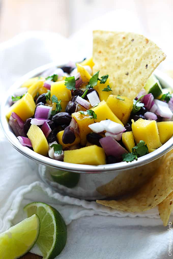 mango black bean salsa in a bowl with a chip dipped in it with slices of limes and chips on the side.