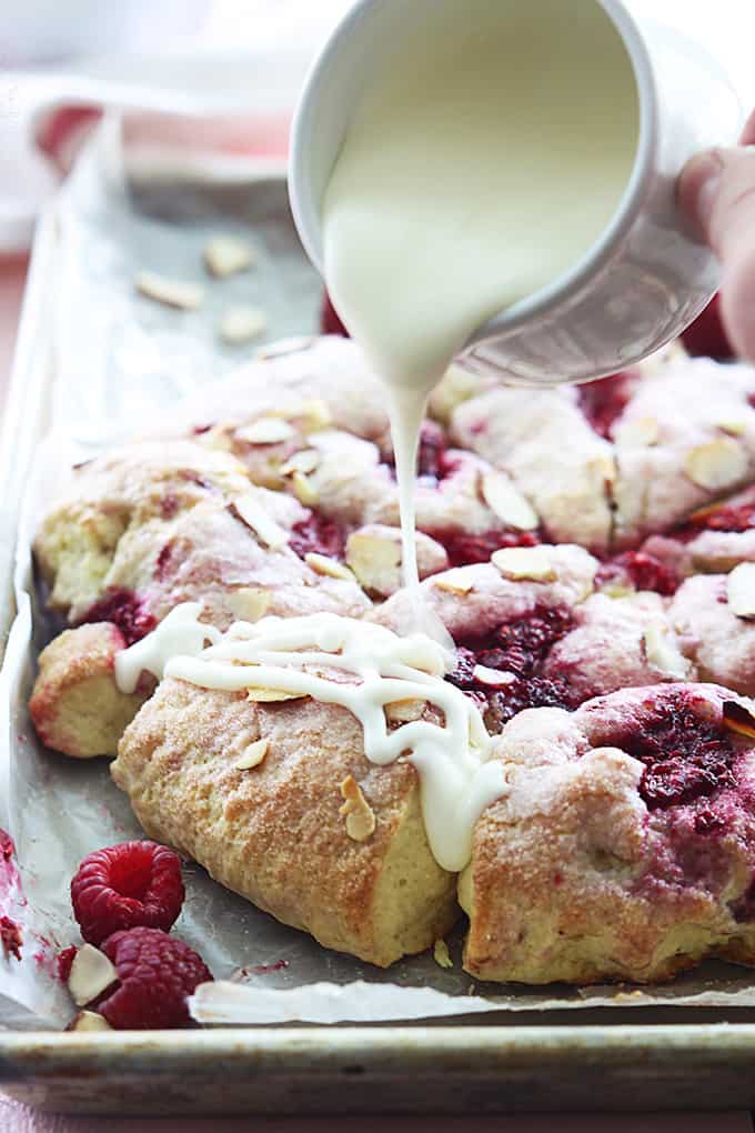 raspberry almond scones on a baking sheet with a hand pouring a cup of almond glaze on top of the scones.