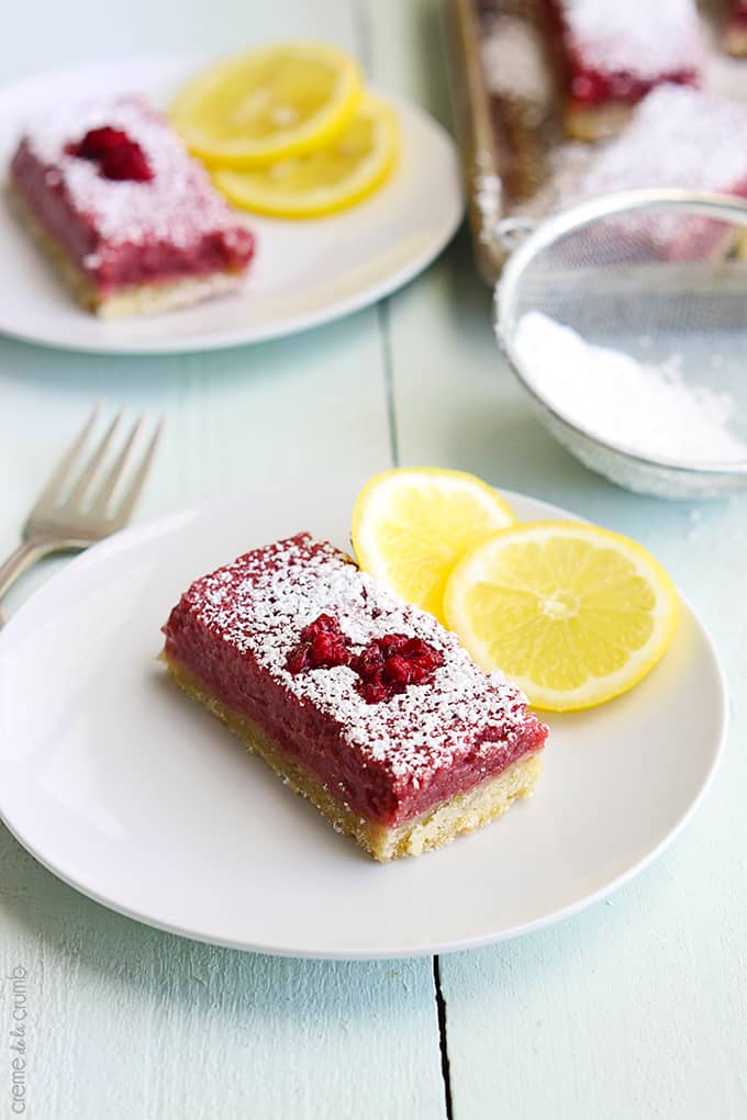  two raspberry lemonade bars on individual plates with slices of lemon on the side with a fork on the table.