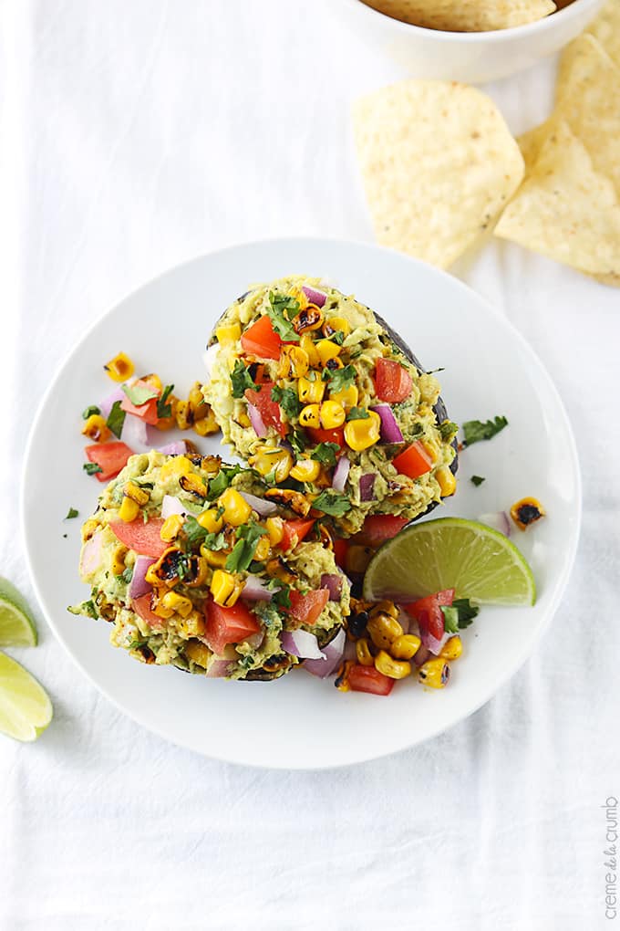 top view of roasted corn fiesta guacamole in hallowed avocados with a slice of a lime on a plate with chips on the side.