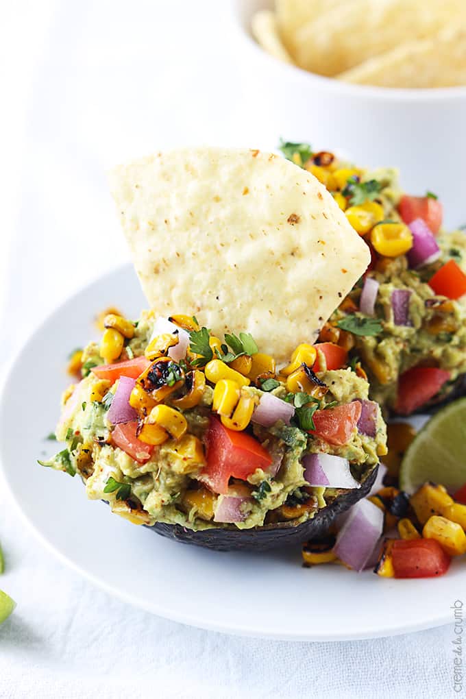 roasted corn fiesta guacamole in hallowed avocados on a plate with a chip dipped in one of the avocados.