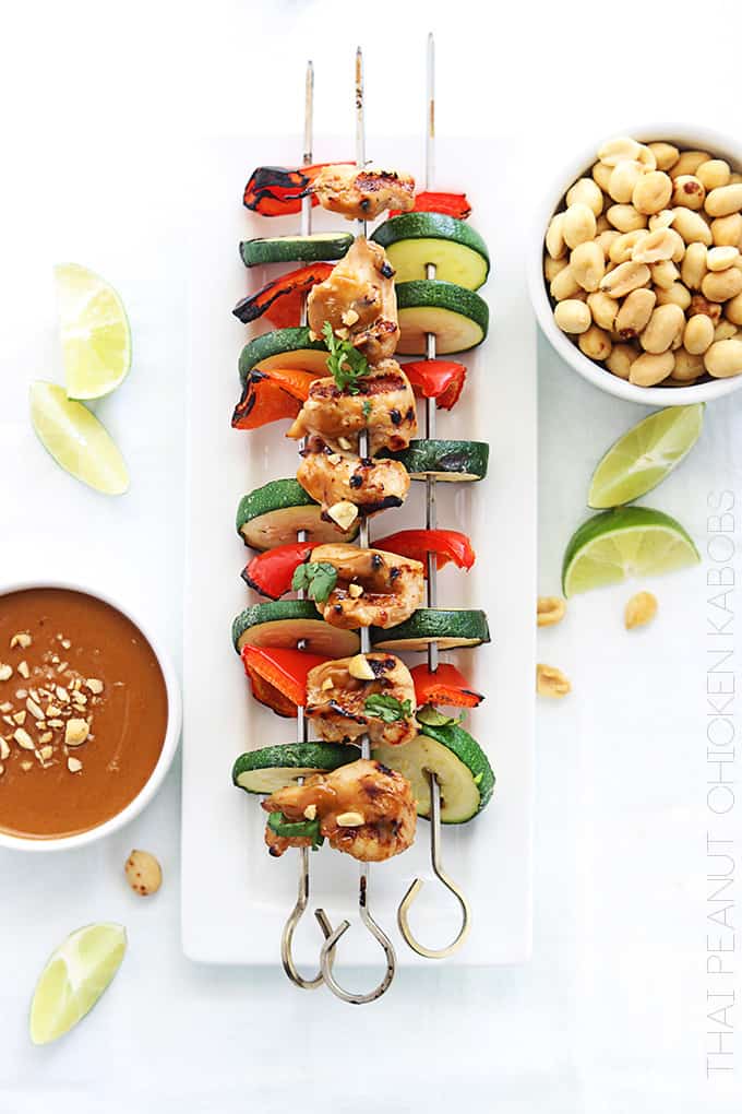 top view of Thai peanut chicken kabobs on a plate with peanut sauce, slices of limes, and peanuts on the side with the title of the recipe written vertically on the bottom right corner going up.