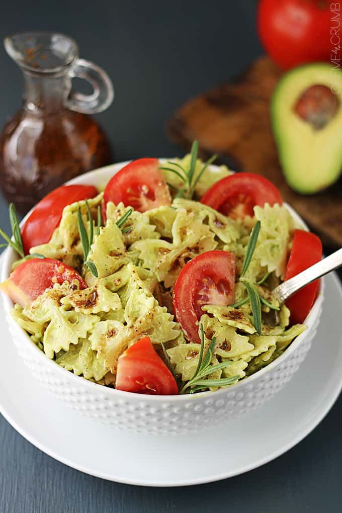 a bowl of creamy avocado pasta with a spoon on a plate with balsamic dressing, a tomato, and half of an avocado faded in the background.
