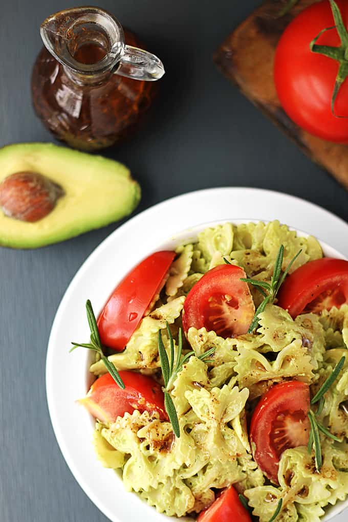 top view of a bowl of creamy avocado pasta with a tomato, half an avocado, and balsamic salad dressing on the side.
