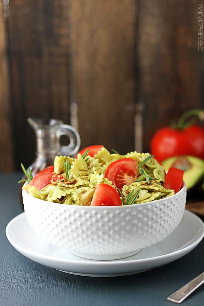 side view of a bowl of creamy avocado pasta on a plate with a spoon on the side and toppings faded in the background.
