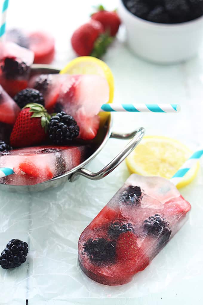 a berry lemonade popsicle with more popsicles in a metal bowl with berries and slices of lemon.
