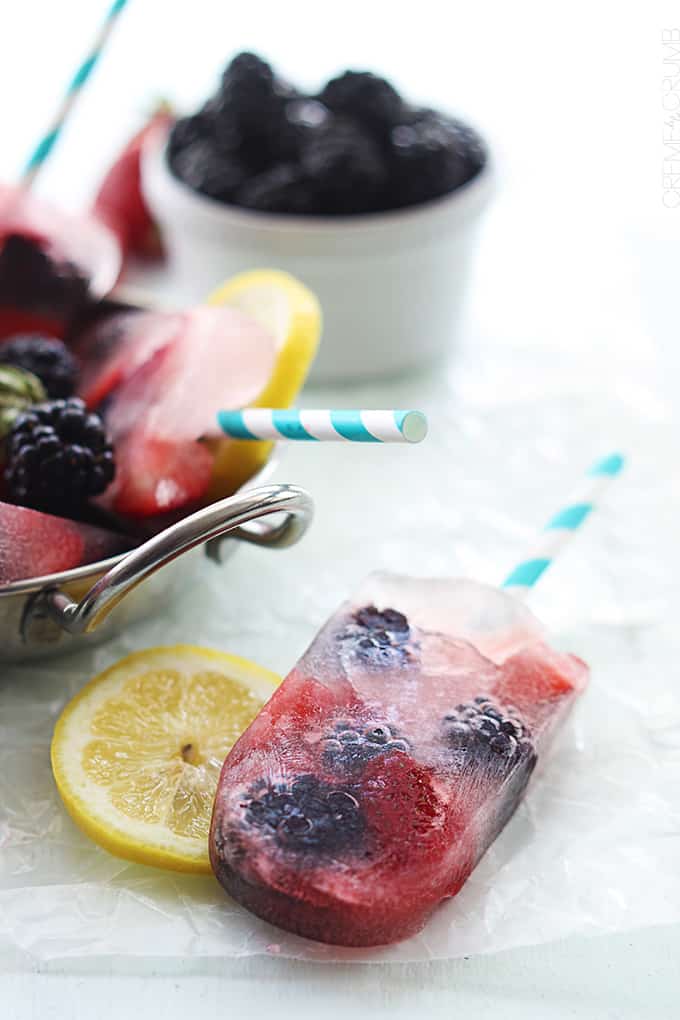 close up of a berry lemonade popsicle on a slice of lemon with a metal bowl full of more popsicles and berries on the side. 