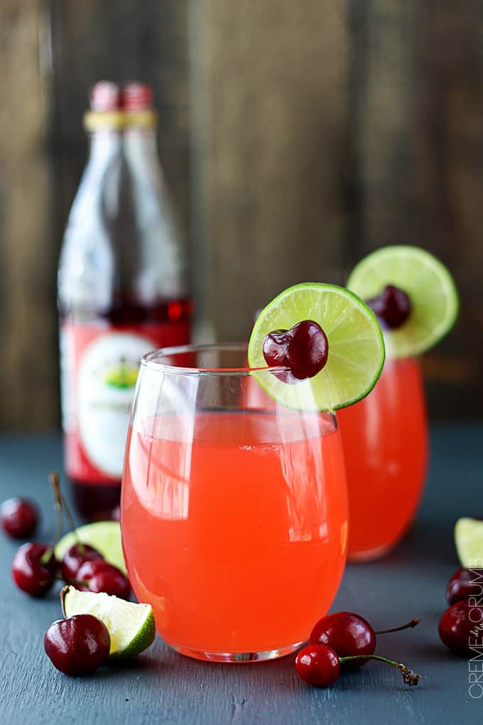 a couple of glasses of cherry limeade with a cherry and a slice of lime on top surrounded by cherries and lime slices with a bottle of Rose's grenadine syrup in the background.