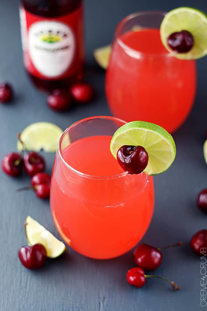 a couple of glasses of cherry limeade with a cherry and a slice of lime on top surrounded by cherries and lime slices.