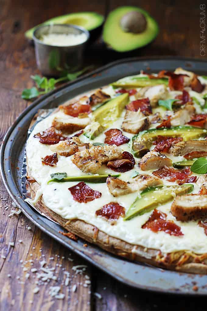 chicken alfredo bacon & avocado pizza in a pizza tray with a small bowl of cheese and two avocado halves in the background.