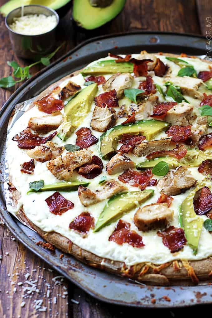 chicken alfredo bacon & avocado pizza on a pizza tray with cheese and two halves of an avocado in the background.