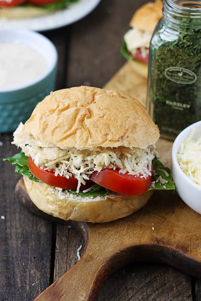 close up of a slow cooker chicken caesar slider on a wooden cutting board with ingredients and more sliders faded in the background