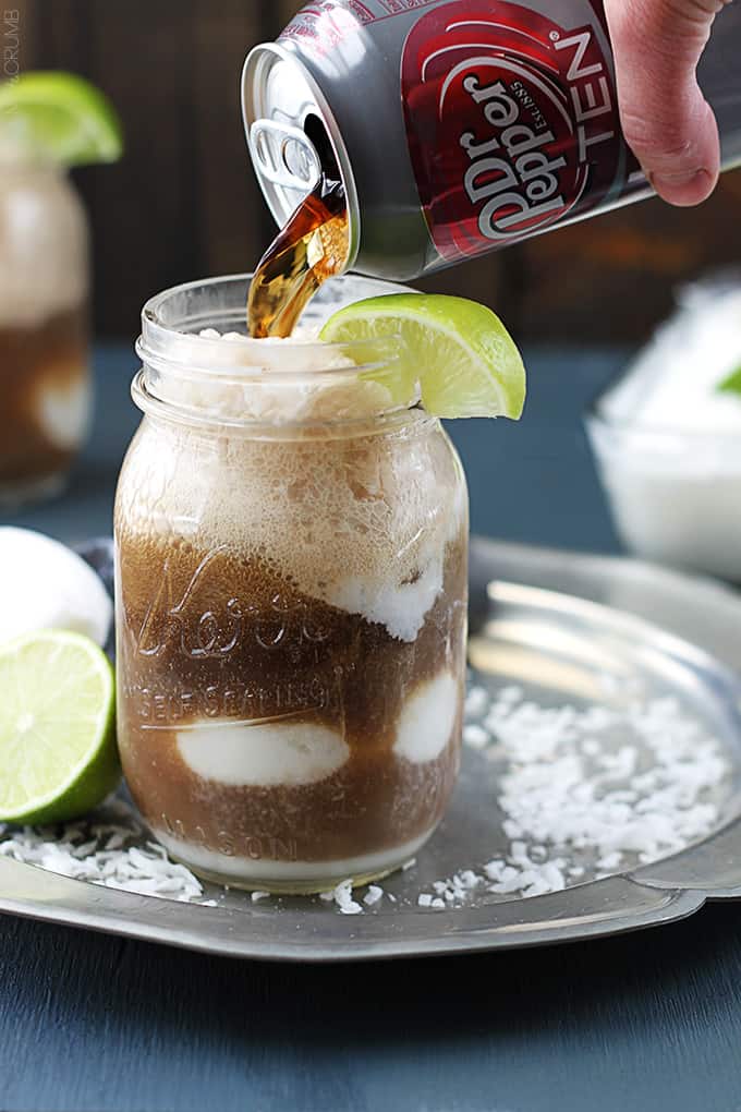 a mason jar full of a dirty Dr. Pepper float with a lime on the top and a hand pouring a can of Dr. Pepper in the mason jar.