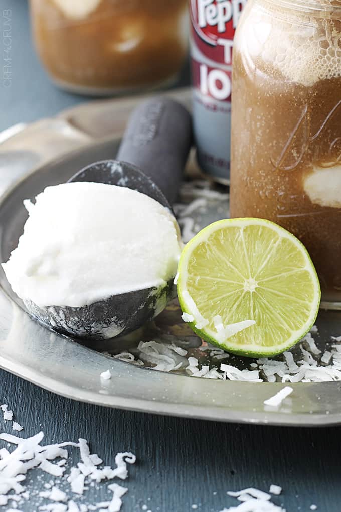 close up of a half of a lime and a ice cream scooper with a scoop of vanilla ice cream on it surrounded by coconut shavings.