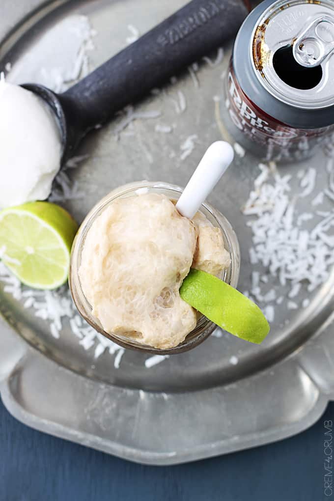 top view of a metal serving tray holding a mason jar of a dirty Dr. Pepper float, a can of Dr. Pepper, half of a lime, and an ice cream scooper with vanilla ice cream on it all surrounded with coconut shavings.