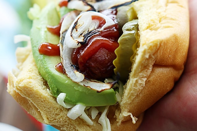 close up of a bbq hot dog with avocado & grilled onions with a side of chips on a lunch tray.