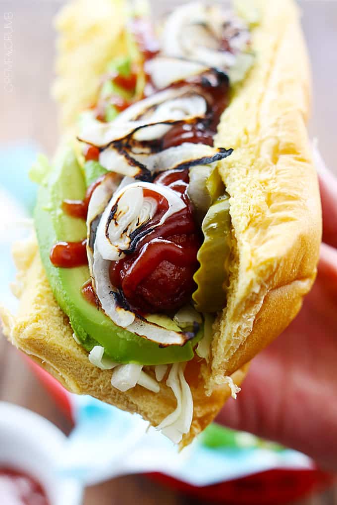 a hand holding bbq hot dog with avocado & grilled onions with a side of chips on a lunch tray.