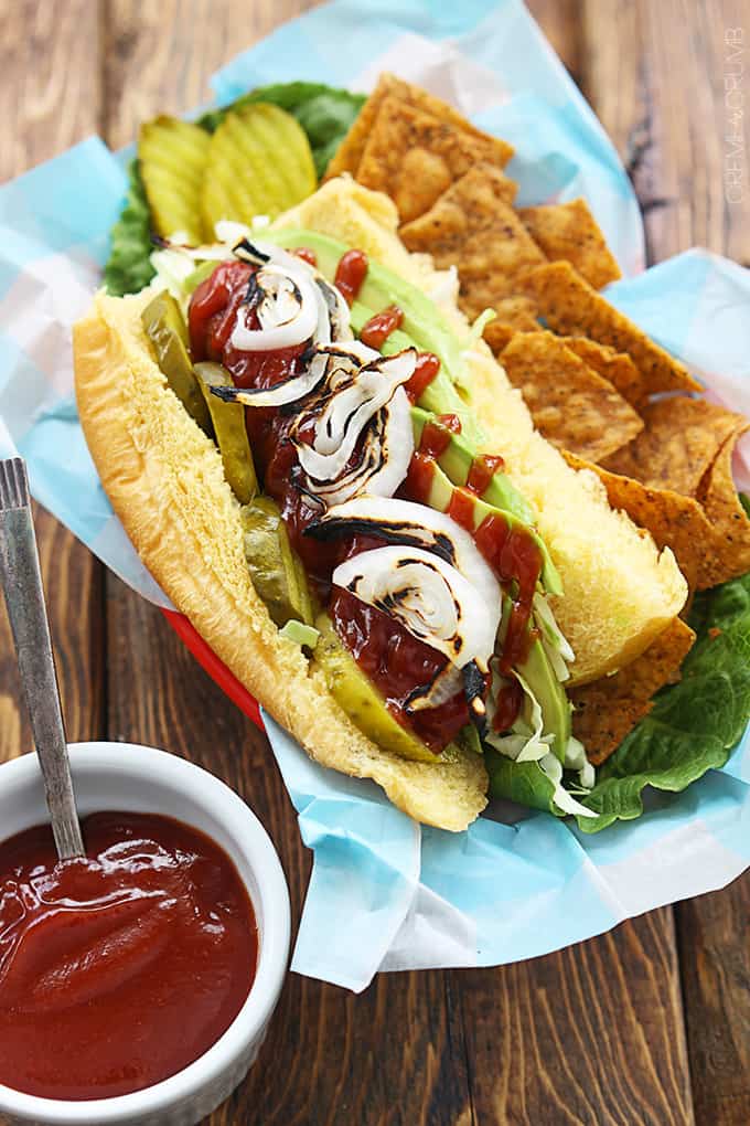 bbq hot dog with avocado & grilled onions with a side of chips on a lunch tray with a side of bbq sauce.