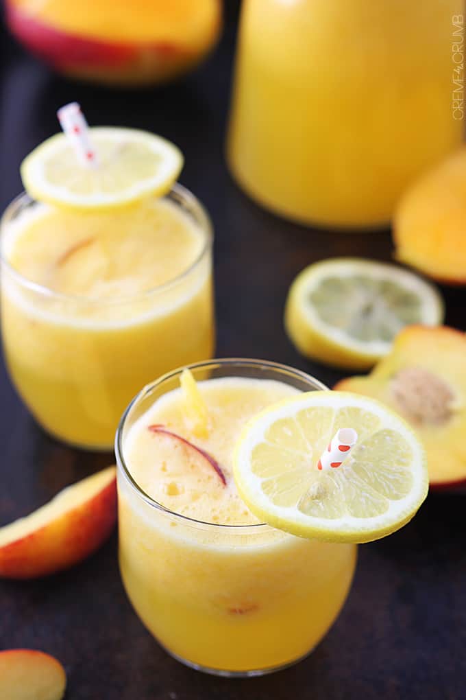 a couple of glasses of mango peach lemonade with slices of lemon on the straws and more slices of lemons and mangos on the side.