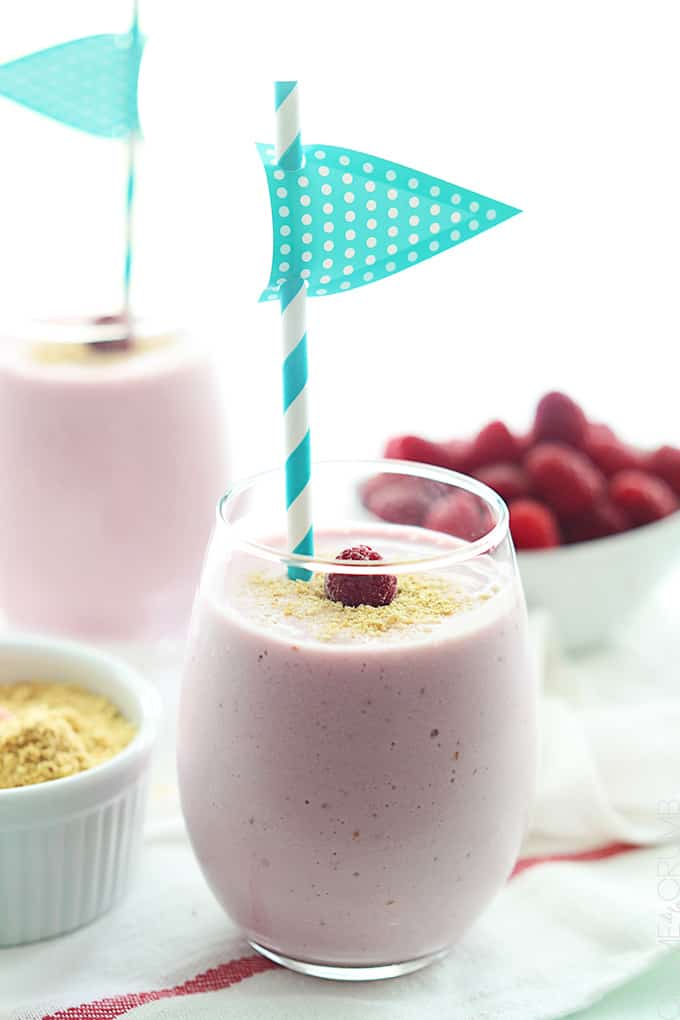a raspberry cheesecake smoothie in a glass with raspberries, crushed graham cracker, and another smoothie faded in the background.