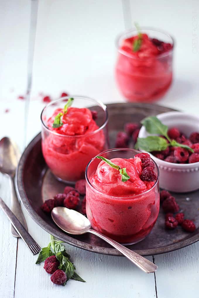 a couple of glasses of raspberry sorbet on a metal plate with spoons and raspberries on the side.